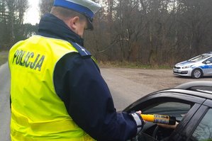 Zdjęcie policjanta Wydziału Ruchu Drogowego, który kontroluje trzeźwość kierowcy samochodu osobowego. Policjant trzyma w ręku urządzenie pomiarowe. Kierowca dmucha do urządzenia przez otwartą szybę pojazdu. W tle widoczny policyjny radiowóz