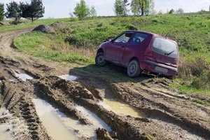 Zdjęcie rozjeżdżonej polnej drogi w dołami wypełnionymi wodą oraz bordowego Fiata Seicento, który zjechał do rowu.