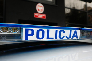 Zdjęcie świetlnego oznakowania policyjnego radiowozu. Wykadrowany niebieski napis &quot;policja&quot; na białym tle. Za radiowozem widoczne wejście do zawierciańskiej komendy w czerwonym godłem i napisem Komenda Powiatowa Policji w Zawierciu.