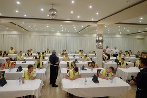 Zawodnicy turnieju w żółtych kamizelkach siedzą na dużej sali przy białych stolikach i piszą test z wiedzy.