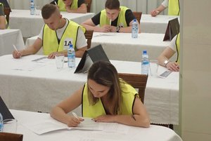 Zawodnicy turnieju w żółtych kamizelkach siedzą na dużej sali przy białych stolikach i piszą test z wiedzy.