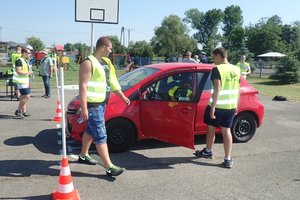 Zawodnicy turnieju w żółtych kamizelkach na torze jazdy sprawnościowej, stojący przy czerwonym samochodzie osobowym.