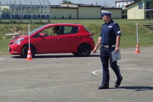 Jadący po torze przeszkód czerwony samochód. Obok stoi policjant oceniający prawidłowe wykonanie zadania.