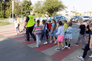 Policjant przeprowadza dzieci przez jezdnie na oznaczonym przejściu dla pieszych.