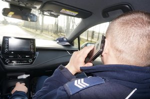 Widoczny policjant rozmawiający przez telefon komórkowy wewnątrz jadącego radiowozu.