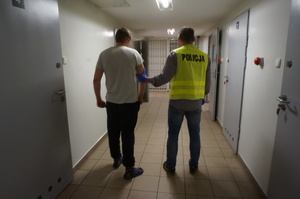 Na korytarzu widoczny policjant w żółtej kamizelce z napisem POLICJA, prowadzi zatrzymanego mężczyznę.