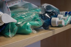Częściowo widoczne: plastikowe butelki z dozownikami z płynem do dezynfekcji oraz zielone kombinezony.