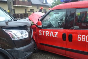 Widoczne uszkodzenbia pojazdów, które zderzyły się ze sobą: czerwonego auta straży pożarnej i brązowego. W tle dwóch strażaków.