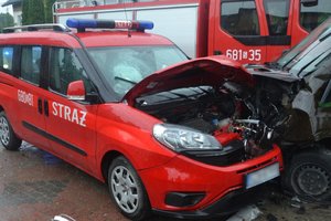 Widoczne uszkodzenbia pojazdów, które zderzyły się ze sobą: czerwonego auta straży pożarnej i brązowego. W tle wóz strażacki.