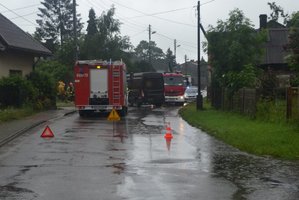 Na pierwszym prlanie dwa pachołki i znak w kształcie trójkąta z wykrzyknikiem i napisem WYPADEK. Widoczne z oddali miejsce zdarzenia. Dwa wozy strażackie, radiowóz oraz brązowy samochód ciężarowy uczestniczący w zderzeniu. Częściowo widoczni strażacy i policjant.