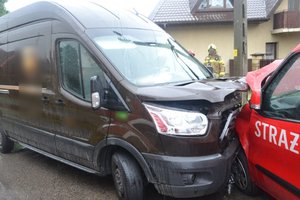 Widoczne dwa pojazdy, które się zderzyły: brązowy ciężarowy i czerwony pojazd straży pożarnej. W tle widoczni strażacy.