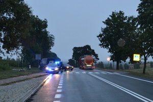 Miejsce zdarzenia drogowego. Widoczne oznakowane przejście dla pieszych, w którego rejonie zaparkowany jest radiowzóz z włączonymi sygnałami świetlnymi oraz pojazd cieżarowy. Przy radiowozie stoi umundurowany policjant. Widoczne także dwa jadące pojazdy.