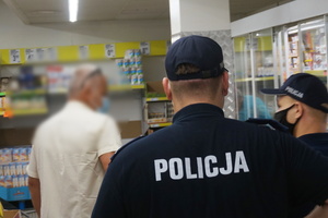 Na zdjęciu widoczni policjanci podczas kontroli przestrzegania obowiązku zakrywania nosa i ust w przestrzeni publicznej. W rozmytym tle widoczny po lewej stronie mężczyzna w  maseczce na twarzy.