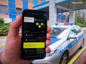 Widoczny trzymany w rękach smartfon z uruchomioną palikacją. W tle widoczny radiowóz i budynek Komendy Wojewódzkiej Policji w Katowicach.