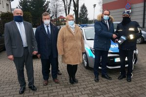 Prezydent Łaz Maciej Kaczyński wraz z Przeowdniczącą Rady Gminy Łazy Anną Staniaszek, a także dwoma radnymi wręczają Komendantowi Komisariatu Policji w Łazach nadkomisarzowi Grzegorzowi Migdałowi kluczyki do nowego, oznakowanego radiowozu. Za nimi widoczny pojazd służbowy.