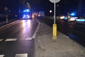 Na zdjęciu miejsce wypadku. Po prawej stronie jezdni widoczny umundurowany policjant, za którym zaparkowane są radiowozy z włączonymi sygnałami świetlnymi.Po lewej stronie jezdni, na przeciwnym pasie widoczny jest wóz strażacki z włączonymi sygnałami świetlnymi. W tle widać jadące pojazdy. Na środku zdjęcia częściowo widać pionowny znak drogowy, usytuowany na wysepce na przejściu dla pieszych.