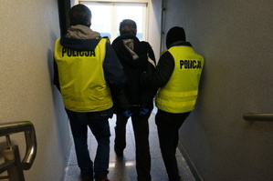 Dwóch policjantów w odblaskowych kamizelkach z napisem POLICJA, prowadzi mężczyznę, który na ręce trzymane z tyłu ma założone kajdanki.