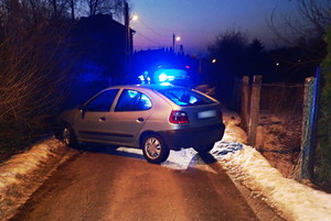 Na zdjęciu widoczny samochód, stojący w poprzek drogi. Za nim widoczny radiowóz z włączonymi sygnałami błyskowymi, a także stojący obok policjant.
