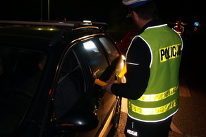Na zdjęciu widoczny umundurowany policjant, który stoi przy samochodzie i trzyma w ręku urządzenie do badania stanu trzeźwości. Za kierownicą częściowo widoczny mężczyzna.