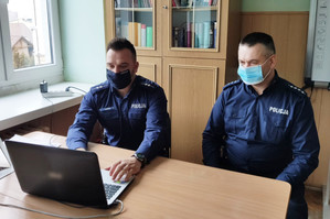 Dwaj umundurowani policjanci z maszeczkami zasłaniającymi usta i nos, siedzą przy biurku, na którym znajduje się laptop i prowadzą prelekcję on-line.