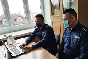 Dwaj umundurowani policjanci z maszeczkami zasłaniającymi usta i nos, siedzą przy biurku, na którym znajduje się laptop i prowadzą prelekcję on-line.