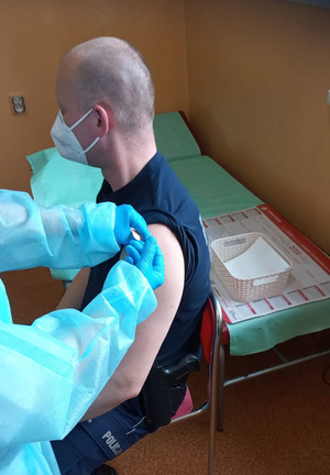 Na zdjęciu widoczny policjant z podciągniętym rękawem lewej ręki, w którą podawana jest mu szczepionka