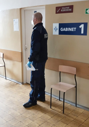 Umundurowany policjant czeka na wejście do gabinetu lekarskiego. Na ścianie widoczny napis GABINET 1