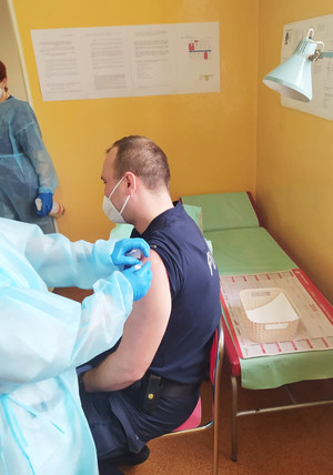 Na zdjęciu widoczny policjant z podciągniętym rękawem lewej ręki, w którą podawana jest mu szczepionka