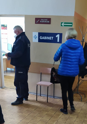 Na zdjęciu widczony umundurowany policjant, czekający na wejście do gabinetu lekarskiego. Za nim w kolejce czeka kobieta.