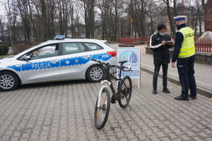 Na zdjęciu widoczny umundurowany policjant podczas badania stanu trzeźwości rowerzysty. Po lewej stronie widoczny rower oraz radiowóz.