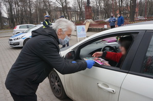 Na zdjęciuw wiodczny mężczyzna w maseczce na ustach i nosie, a także w rękawiczkach jednorazowych, który przekazuje kierującej samochodem kobiecie ulotki i długopis. W tle widać umundurowanych policjantów, radiowóz oraz dwie stojące osoby.