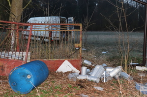 Na zdjęciu widoczne leżące na ziemi aluminiowe rury. Po lewej stronie przewrócona na ziemię niebieska beczka. Za nimi widoczne przęsła bramy. W tle na ziemi leżące alumionowe rury prowadzące, do widocznego białego samochodu dostawczego z otwartymi prawymi tylnymi drzwiami.