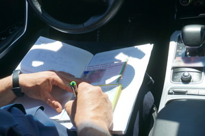 Na zdjęciu widoczny mandat karny, wypisywany przez policjanta oraz dokumenty kierowcy. Dane rozmyte.