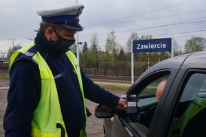 Umundurowany policjant w kamizelce doblaskowej pokazuje kierowcy na alcosensorze wynik badania stanu trzeźwości. W tle widoczna tablica z napsiem: Zawiercie