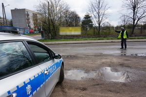 Na zdjęciu widoczny częściowo radiowóz i umundurowany policjant w kamizelce odblaskowej, dający znak do zatrzymania pojazdu.