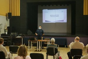 Umundurowany policjant prowadzi prelekcję dla seniorów. Przed nim widoczne siedzące na krzesłach osoby.