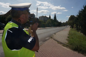 Na zdjęciu widoczny umundurowany policjant mierzący prędkość nadjeżdżającego pojazdu.