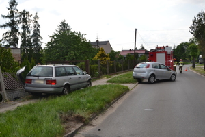 Na zdjęciu widoczne dwa samochody, które brały udział w zdarzeniu drogowym. Jeden wjechał w płot, drui stoi na krawężniku i na jezdni. W tle widoczny wóz strażacki i strażacy.