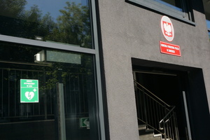 Na zdjęciu widoczne główne wejście do budynku Komendy Powiatowej Policji w Zawierciu, po którego lewej stronie widoczna jest zielona, odblaskowa tabliczka z napisem: Obiekt wyposażony w AED oraz grafiką przedstawiającą serce z błyskawicą i plusem
