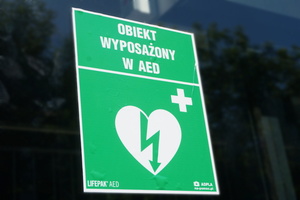 Na zdjęciu widoczna zielona, odblaskowa tabliczka z grafiką przedstawiającą serce z błyskaiwcą i plusem po prawej stronie. Nad nią widoczny napis: Obiekty wyposażony w AED