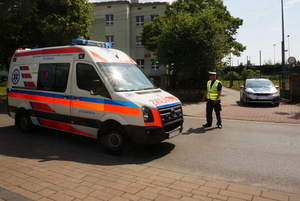 Na zdjęciu z lewej strony widoczny jadący ambulans. Na jezdni stoi umundurowany policjant. W tle widoczny radiowóz.