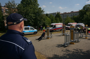 Na pierwszym planie, w lewym doplnym rogu widoczny częściowo umundurowany policjant. W tle widoczny radiowóz oraz trzy ambulans. W centralnej cześci widoczny siedzący na sprzęcie do ćwiczeń mężczyzna.