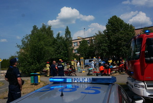 Na zdjęciu po lewej stronie widoczny umundurowany policjant, w centralnej części radiowóz z włączonymi sygnałami świetlnymi. Po prawej stronie częściowo widoczny wóz strażacki. W tle widoczni ratownicy i strażacy udzielający pomocy osobom oraz umundurowany policjant stojący obok mężczyzny.