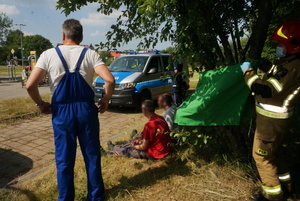 Na zdjęciu widoczni trzej mężczyźni. Jeden z nich stoi, dwóch siedzi na trawie. Obok strażak na drzewie zawiesza zieloną płachtę, oznaczając miejsce lokowania poszkodowanych określonych w traiżu, jako &quot;zieloni&quot;, czyli chodzący. W tle widoczny radiowóz i stojący obok niego umundurowany policjant.