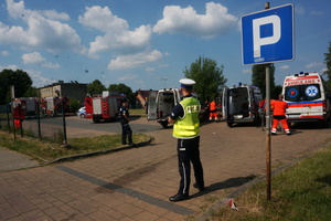 Na zdjęciu widoczni dwaj umundurowani policjanci zabezpieczający wjazd na teren parkingu. W tle widoczne wozy strażackie oraz ambulanse, a także ratownicy udzielający pomocy.