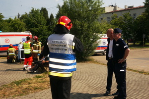 Na zdjęciu widoczny strażak w kamizelce z napisem: Kierujący działaniem ratowniczym. Obok niego stoi umundurowany policjant i mężczyzna w garniturze. W tle widoczne działania ratunkowe prowadzone przez ratowników i strażaków. Widoczne także ambulanse.