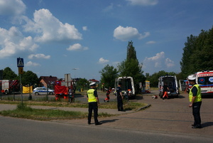 Na zdjęciu widoczni trzej umundurowani policjanci zabezpieczający wjazd na teren parkingu. W tle widoczny radiowóz, wozy strażackie, a także ambulanse. Widoczni także ratownicy.