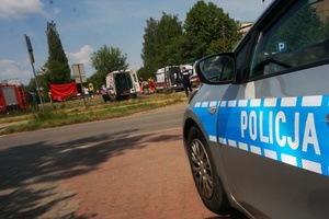 Na zdjęciu widoczny częściowo radiowóz, w tle wóz strażacki i ambulanse, a także ratownicy prowadzący akcję ratunkową.
