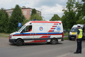Na zdjęciu widoczny jadący ambulans, za którym jedzie inny dostawczy samochód oraz stojący na jezdni policjant, kierujący ruchem.