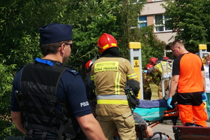 Na zdjęciu widoczny umundurowany policjant, w tle udzielający pomocy strażacy i ratownicy.